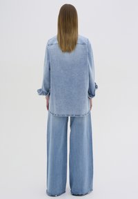 Lyseblå denimjakke med lange ærmer og buet kant, parret med brede lyseblå denimbukser. Begge genstande har synlige sømme.