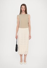 Débardeur beige clair sans manches avec un détail unique aux épaules, associé à une jupe midi en dentelle ivoire comportant des motifs en découpes circulaires. Sac à main noir.