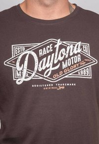 T-shirt marron en coton, avec un motif graphique vintage en texte blanc et orange portant les inscriptions "Daytona Motor Supply" et "1989."