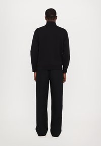 Pullover nero con collo alto e maniche lunghe, abbinato a pantaloni neri a gamba larga e sneaker bianche, visto da dietro.