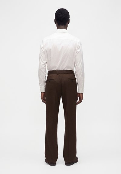 Homme portant une chemise blanche à manches longues et un pantalon marron, debout de dos devant un fond clair uni.