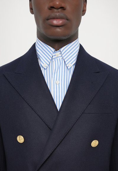 Blazer bleu marine croisé avec des boutons dorés sur une chemise à rayures bleu clair et blanche. Tissu lisse, coupe ajustée, sans motifs visibles.