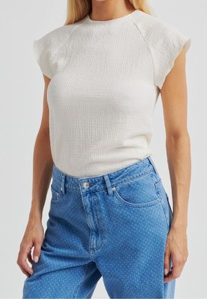 Femme portant un haut blanc texturé à manches courtes associé à un jean bleu taille haute avec un motif discret à pois.
