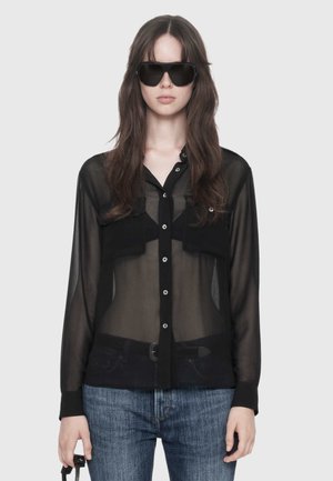 Woman wearing large black sunglasses, sheer black button-up shirt over a black bra, and blue jeans, holding a black handbag.