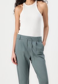 Weißes, geripptes Tanktop kombiniert mit hellgrünen, locker sitzenden Hosen mit Kordelzug und seitlichen Taschen. Glatte Stofftextur.