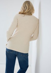 Vrouw met blond haar leunt tegen een witte muur, draagt een beige trui en blauwe spijkerbroek, met haar gezicht van de camera afgekeerd.