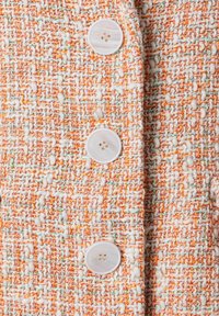 Tissu texturé avec des fils multicolores en orange, blanc et vert. Présente de grands boutons ronds de couleur claire le long du devant.