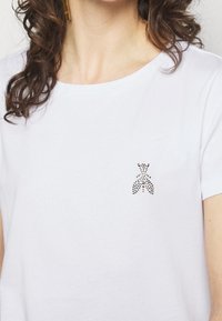 Bílá bavlněná tričko s kulatým výstřihem, s malým zdobeným designem černých a stříbrných kamínků na levé části hrudi.