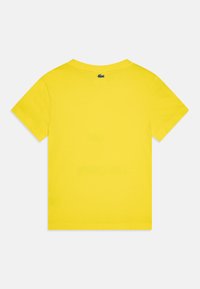 T-shirt en coton jaune, à manches courtes, col rond. Présente un petit logo vert au-dessus du col. Texture lisse, coupe décontractée.