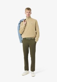 Maglione beige con zip al collo, pantaloni verde oliva, scarpe da ginnastica bianche. Giacca di jeans chiara appoggiata sulla spalla. Accento verde con logo sul maglione.