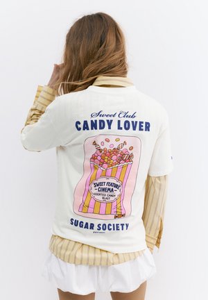Personne aux longs cheveux portant une chemise blanche par-dessus un t-shirt jaune rayé, avec un motif coloré d’un sac de bonbons et le texte « Candy Lover » dans le dos.