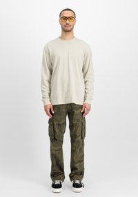 Beige shirt met lange mouwen, olijfgroene camouflage cargobroek met zakken en zwarte sneakers. Gele getinte zonnebril maakt de look compleet.