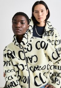 Veste zippée en polaire de couleur crème douce avec un motif de texte noir audacieux. Col haut, coupe décontractée et fermeture éclair à l'avant. Tissu texturé et chaud.