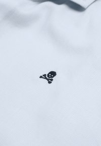 Witte katoenen polo-shirt met een kleine zwarte doodskop en gekruiste botten borduursel aan de linkerkant, textuurstof met fijne ribbels.