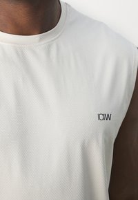 Gros plan d'une personne portant un débardeur blanc de sport avec un petit logo "ICIW" sur la poitrine.