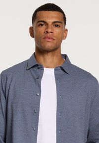 Siva srajca s kragnjo, izdelana iz mehkega materiala, z navadnim ovratnikom, sprednjim zapiranjem na gumbe in belim spodnjim majico vidnim spodaj.