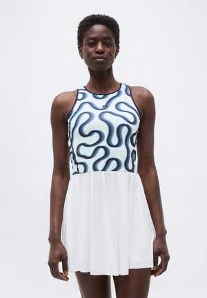 Γυναίκα που φοράει φορεματίνα χωρίς μανίκια με μαύρο αφηρημένο σχέδιο στην κορυφή και απλό λευκό φόρεμα στη φούστα, στέκεται μπροστά σε απλό φόντο.