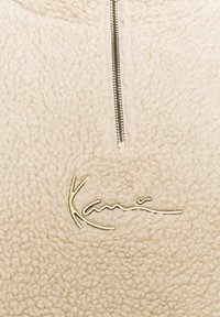 Pullover in pile beige con una zip a quarto sul davanti e un logo ricamato dorato "Kani". Il tessuto è strutturato e morbido, creando un aspetto accogliente.