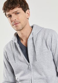 Haut à capuche zippé gris avec cordons, fabriqué dans un matériau doux. Doté d'une fermeture éclair complète et de poches, porté par-dessus un t-shirt col en V sombre.