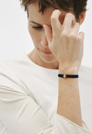 Persona con cabello corto y castaño que lleva una blusa blanca, sosteniéndose la frente con una mano que lleva una pulsera oscura con un charm de estrella dorada.