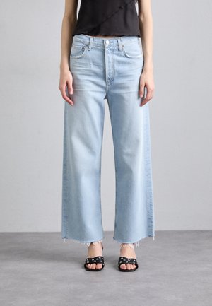 Person iført lyseblå jeans med brede ben og rå kanter, en sort cropped top og sorte åbne sandaler med sølvnitter.