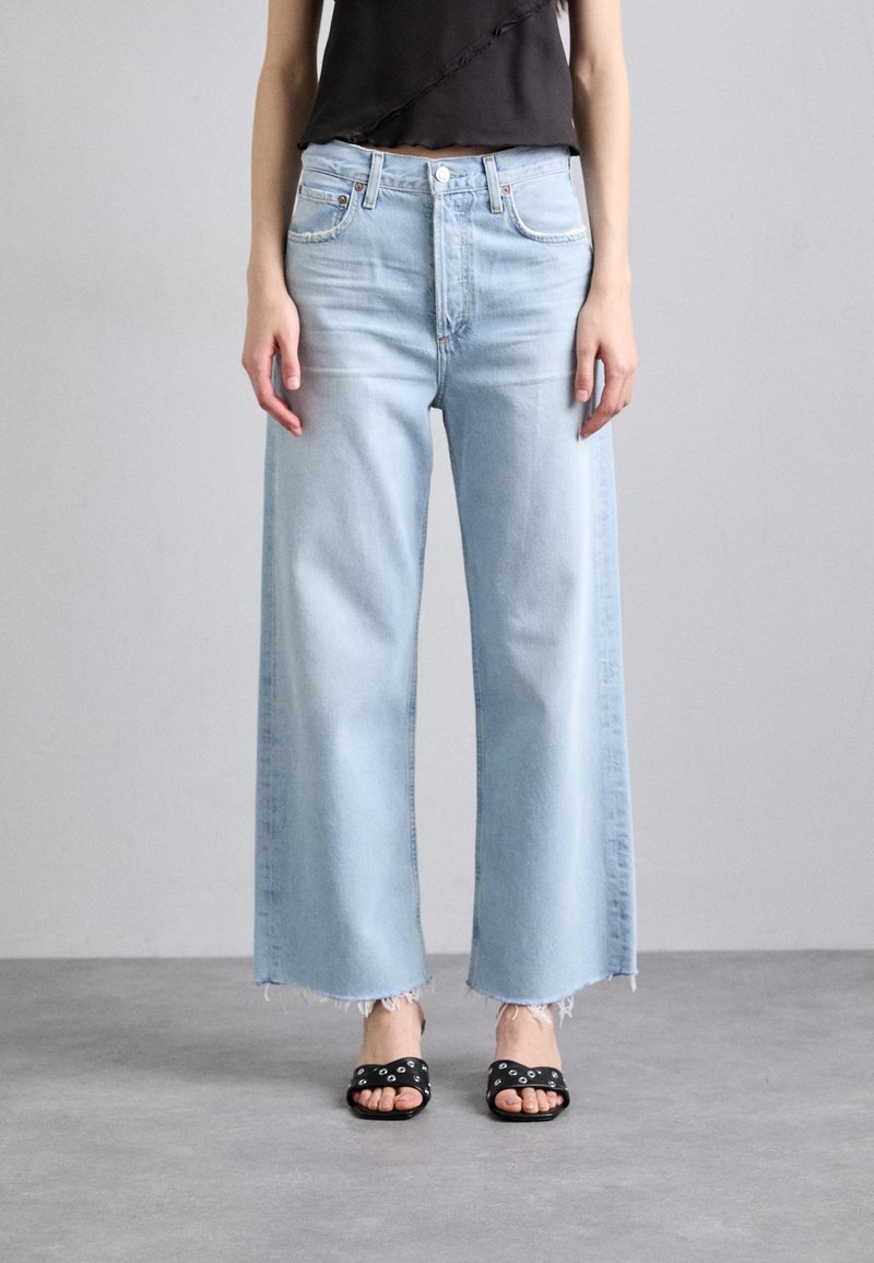 Personne portant un jean bleu clair à jambes larges avec des ourlets bruts, un top court noir et des sandales noires à bout ouvert avec des clous argentés.