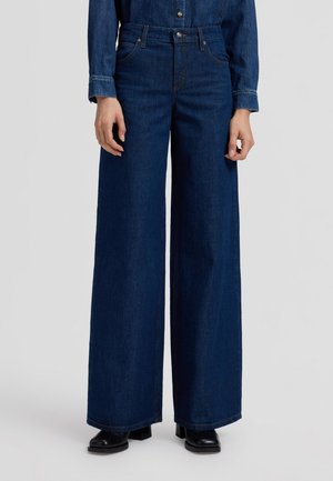 Personne portant un jean large bleu foncé, une chemise en denim assortie et des bottes noires à talons, debout devant un fond uni.