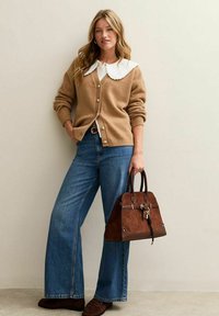 Cárdigan beige con botones dorados sobre una blusa blanca con un gran cuello escalloped. Jeans de pierna ancha azules y un bolso estructurado marrón.