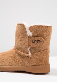 Botte UGG en daim marron avec doublure en polaire blanche visible et semelle en caoutchouc, affichant un patch logo sur le côté sur un fond uni.
