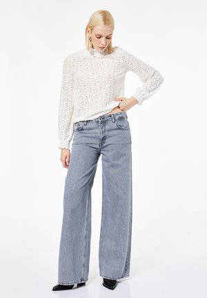 Donna che indossa una camicetta in pizzo bianca a maniche lunghe infilata in jeans grigi a vita alta e gambe larghe, e scarpe nere con punta affusolata, in piedi su sfondo bianco.