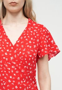 Γυναίκα που φοράει κόκκινο φόρεμα τύπου wrap με μικρό λευκό λουλουδάτο μοτίβο και κοντά μανίκια με βολάν, πάνω σε φωτεινό φόντο.