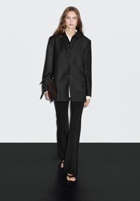 Black oversized blazer with buttons, paired with black tailored pants. Model holds a dark brown clutch. Simple backdrop, minimalistic design.