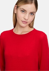 Roter Strickpullover mit langen Ärmeln, runder Ausschnitt und weichem Textur. Der Stoff wirkt warm und leicht strukturiert.