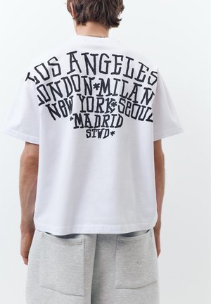 Persoană purtând un tricou alb cu text mare negru care numește orașe: Los Angeles, Londra, Milano, New York, Seul, Madrid, și pantaloni scurți gri.