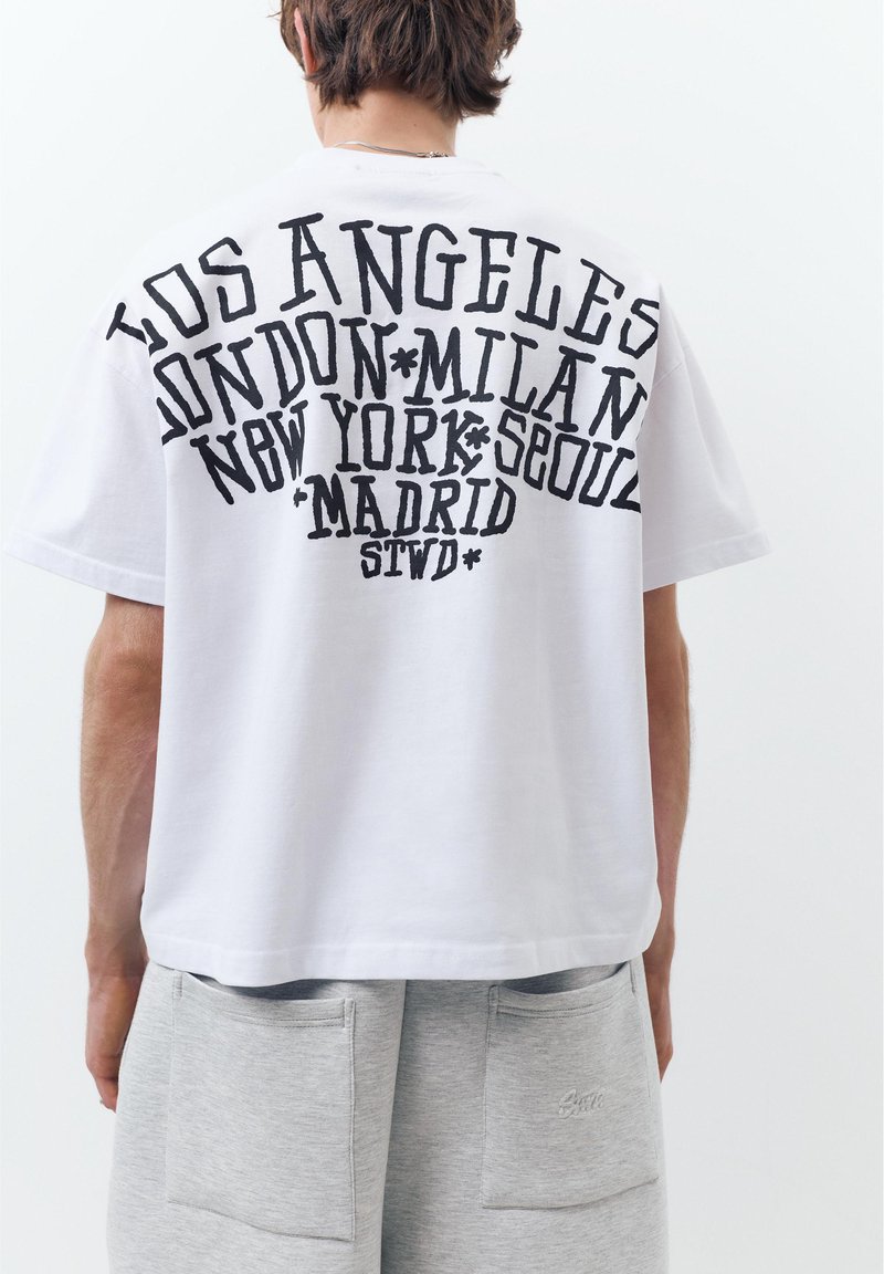 Personne portant un t-shirt blanc avec un grand texte noir listant des villes : Los Angeles, Londres, Milan, New York, Séoul, Madrid, et un short gris.
