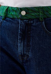 Vaqueros de mezclilla con un lavado azul oscuro, con un cinturón de patrón vibrante en verde y negro, botón de metal visible y costuras naranjas en contraste.