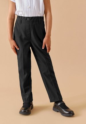 Enfant portant un pantalon droit noir, des chaussures Mary Jane noires et un chemisier blanc à manches courtes, debout sur un fond beige.