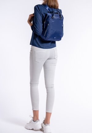 Vrouw in een witte skinny jeans, witte sneakers en een blauw polka-dot shirt, met een marineblauwe rugzak op haar rug.