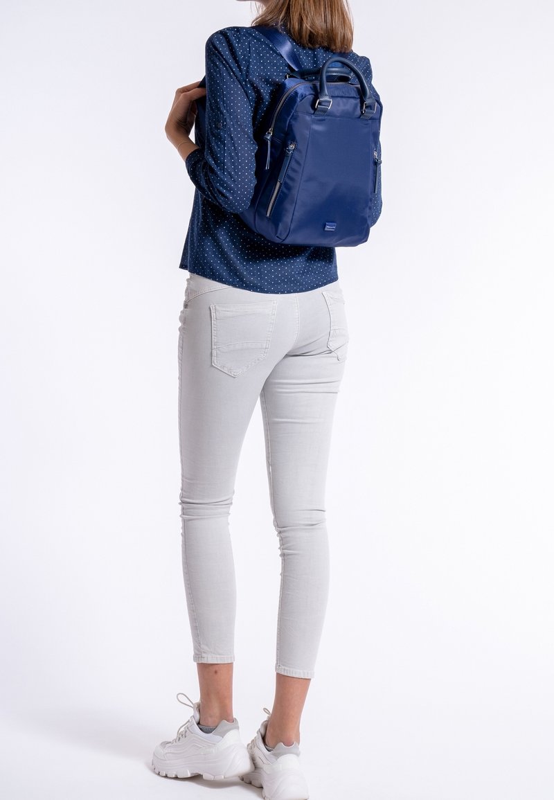 Kvinna iklädd vita skinny jeans, vita sneakers och en blå prickig skjorta, bär en marinblå ryggsäck på ryggen.