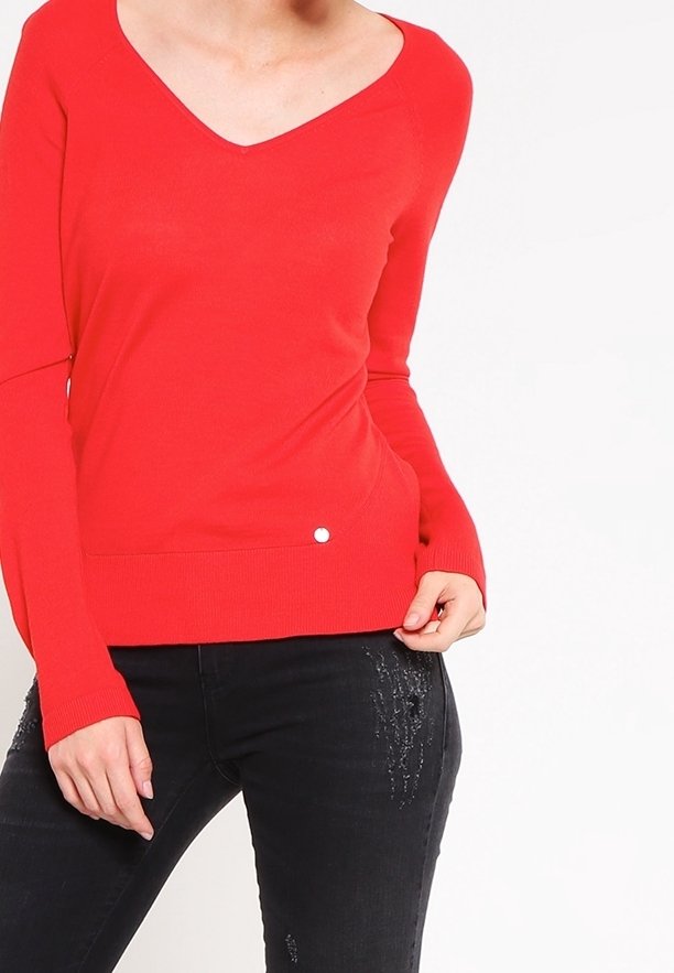 Rode gebreide trui met een V-hals, lange mouwen en een detail met knopen aan de zijkant. Gepaard met zwarte jeans met verfijnde versieringen.