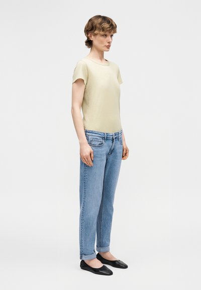 Νεαρό άτομο με μπεζ κοντομάνικο τοπ, ανοιχτό μπλε τζιν με γυρισμένα μπατζάκια και μαύρα φλατ παπούτσια, στέκεται κοιτάζοντας προς τα δεξιά σε ένα απλό λευκό φόντο.