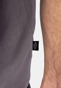 Šedé tričko z látky s bočním štítkem a diamantovým logem. Materiál má hladký, mírně texturovaný povrch.