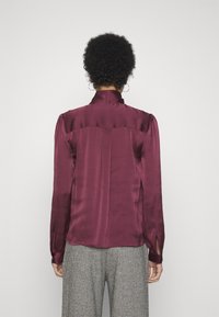 Blusa in satin bordeaux con maniche lunghe, colletto alto e dettagli plissettati sulle spalle. Presenta spacchi laterali all'orlo.