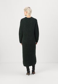 Μακρύ, σκούρο πράσινο, πλεκτό φόρεμα με υφή και μακριά μανίκια, σε ευθεία γραμμή, συμπληρωμένο με μαύρες μπότες έως το γόνατο.