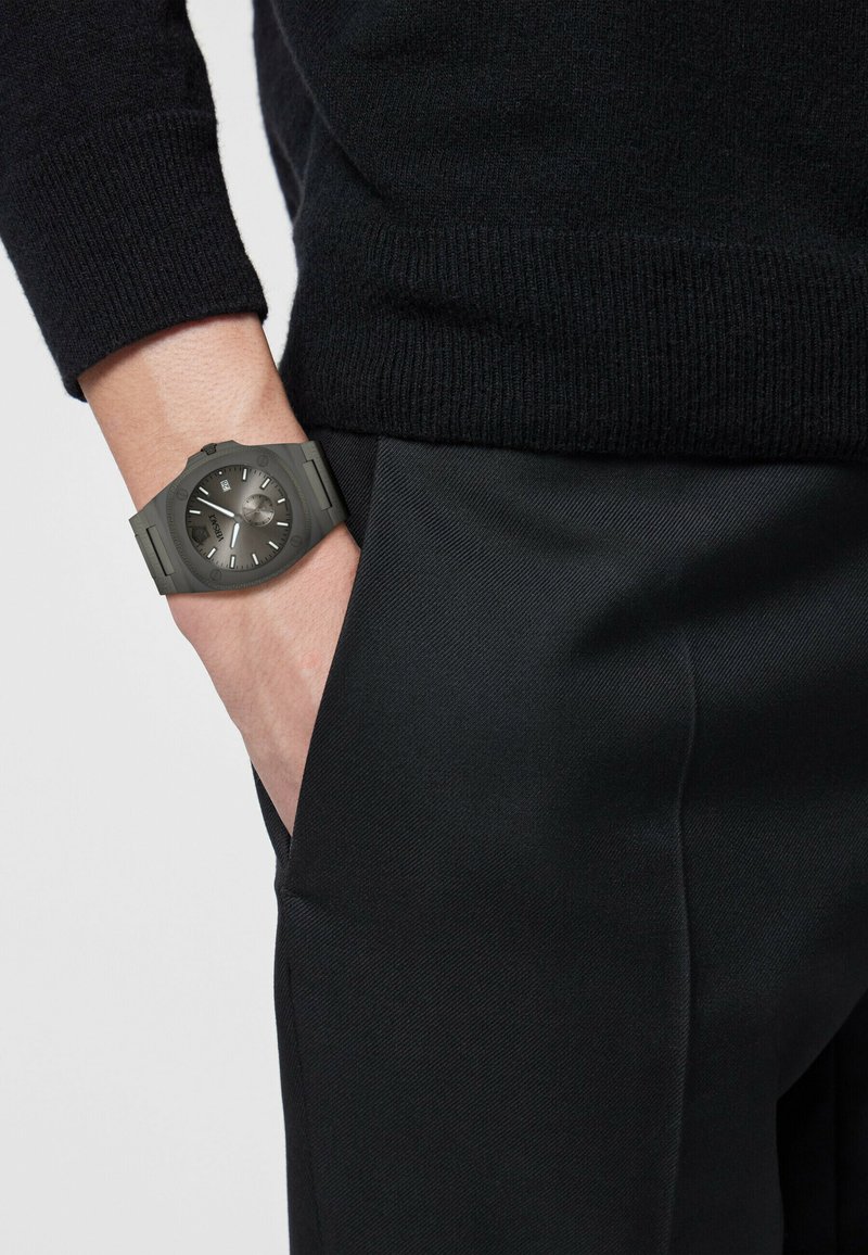 Grijze silicone horloge met ronde wijzerplaat, contrasterende uurmarkeringen, gedragen aan een hand gestoken in donkere broeken, onder een zwarte mouw van een trui.