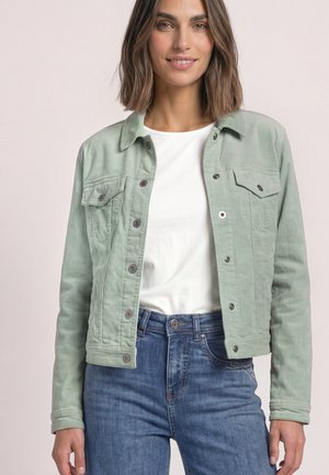 Femme aux cheveux bruns mi-longs portant une veste boutonnée vert clair par-dessus une chemise blanche et un jean bleu, souriant doucement.