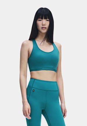 Junge Frau mit langen schwarzen Haaren, die einen tealfarbenen Sport-BH und passende hoch taillierte Leggings vor einem schlichten Hintergrund trägt.
