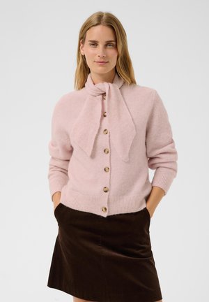 Femme portant un cardigan rose clair boutonné avec un détail foulard noué et une jupe marron foncé, debout les mains dans les poches.