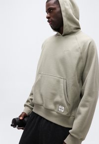 Άνδρας που φοράει μπεζ φούτερ Nike, κρατώντας ένα μαύρο παγούρι, κοιτάει προς τα πλάγια σε ένα λευκό απλό φόντο.