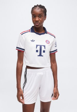 Femme portant un maillot de football blanc raccourci du Bayern Munich avec des bordures bleu marine et rouge et un short blanc assorti avec des rayures noires.